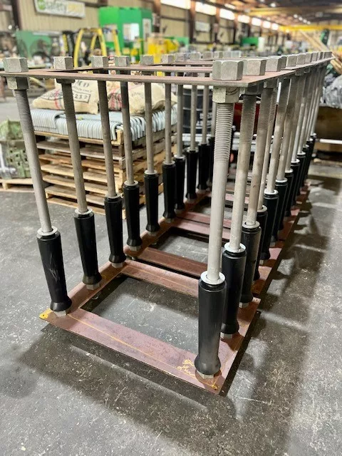 Fabricated anchor bolt assembly with template plates and black bell bottom sleeves on the shop floor.
